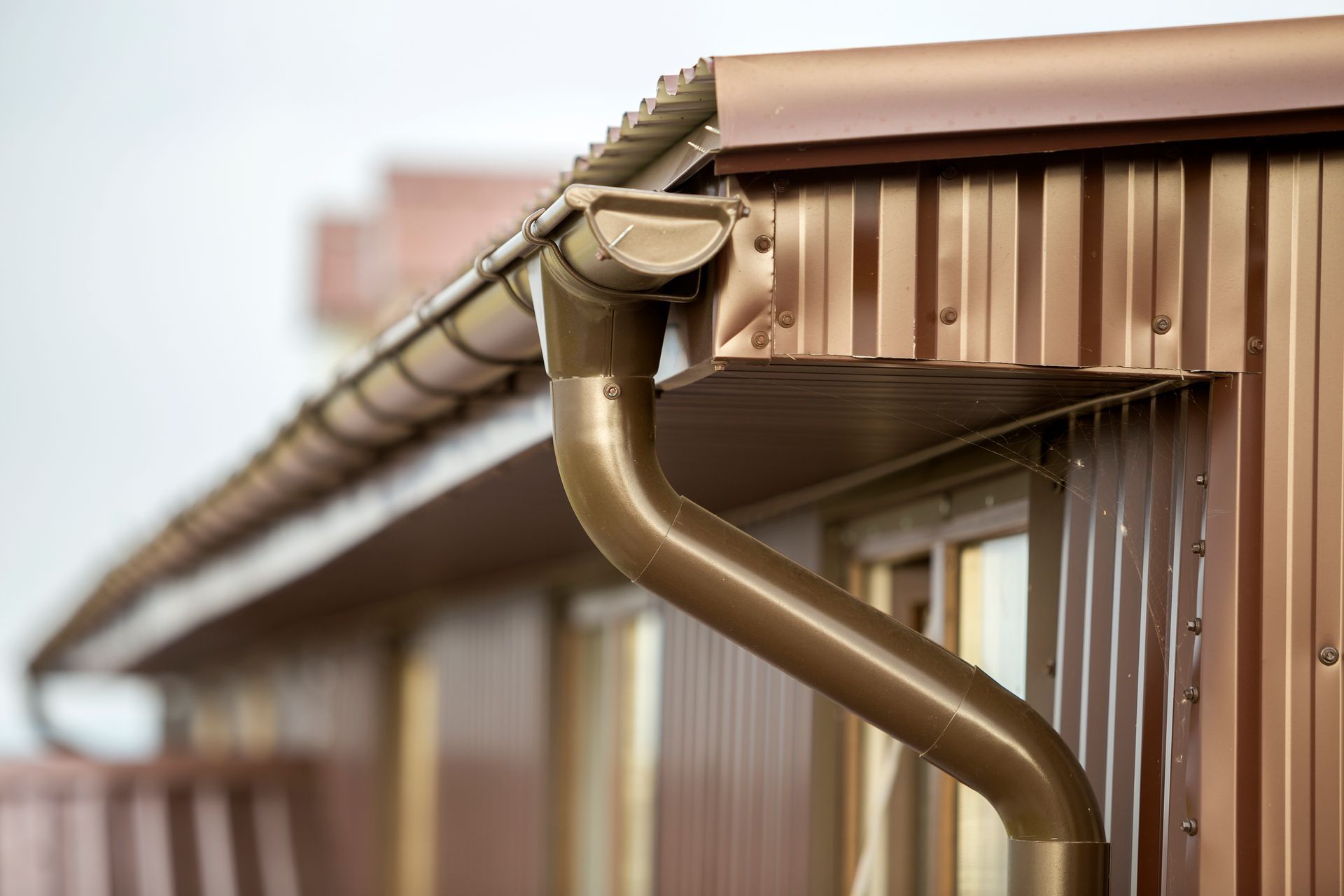 Brown metal rain gutter on the edge of a corrugated roof against a blurred building background