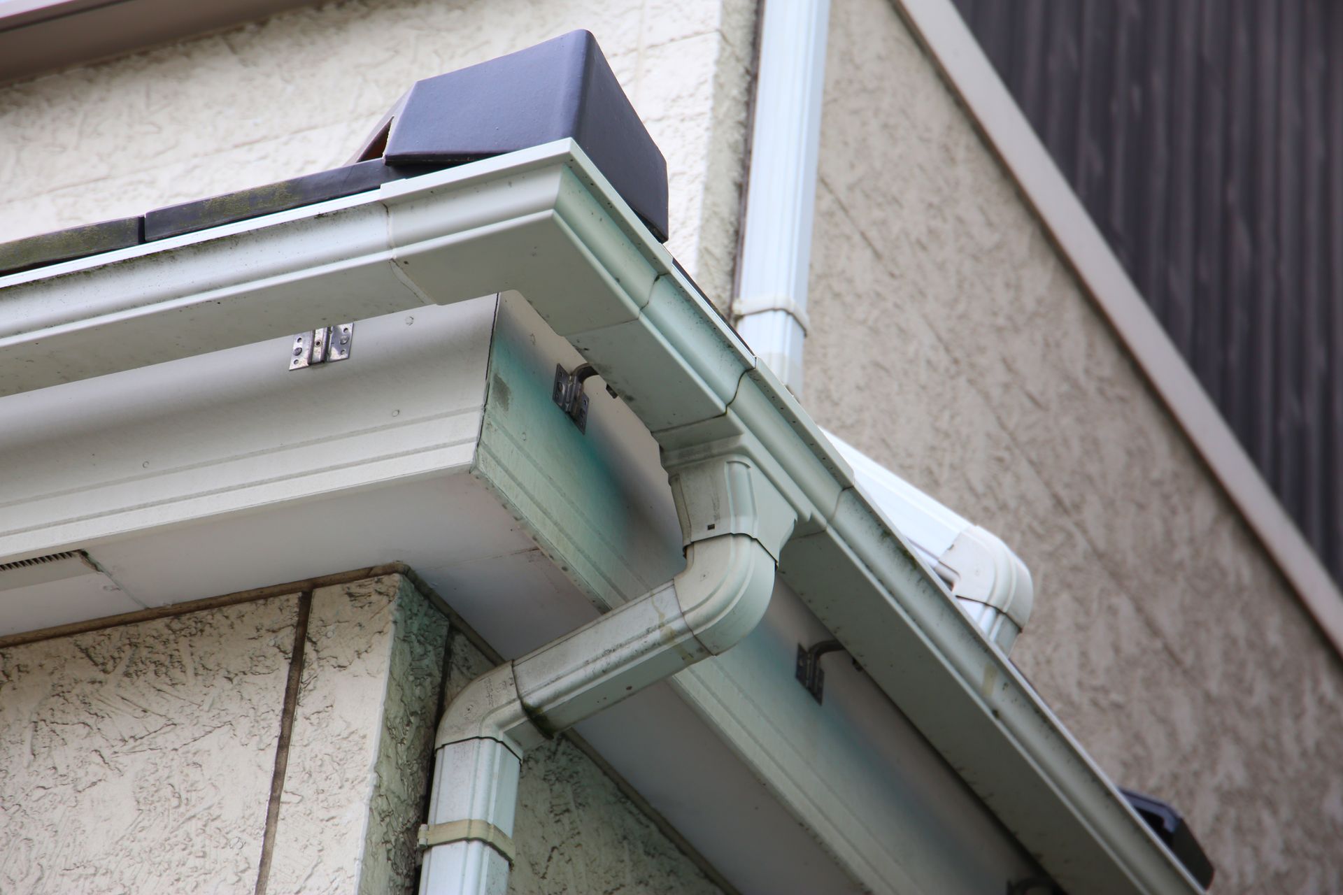 Close-up of house eaves with white gutters and downspout against beige siding