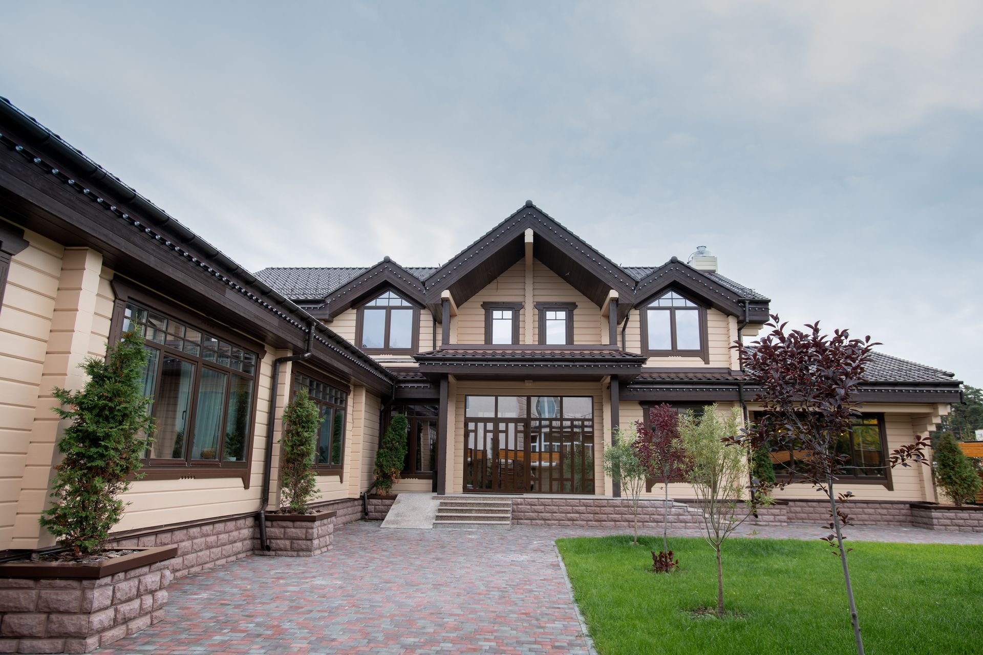 Tan two-story house with dark brown trim, brick facade, and green lawn under a cloudy sky.