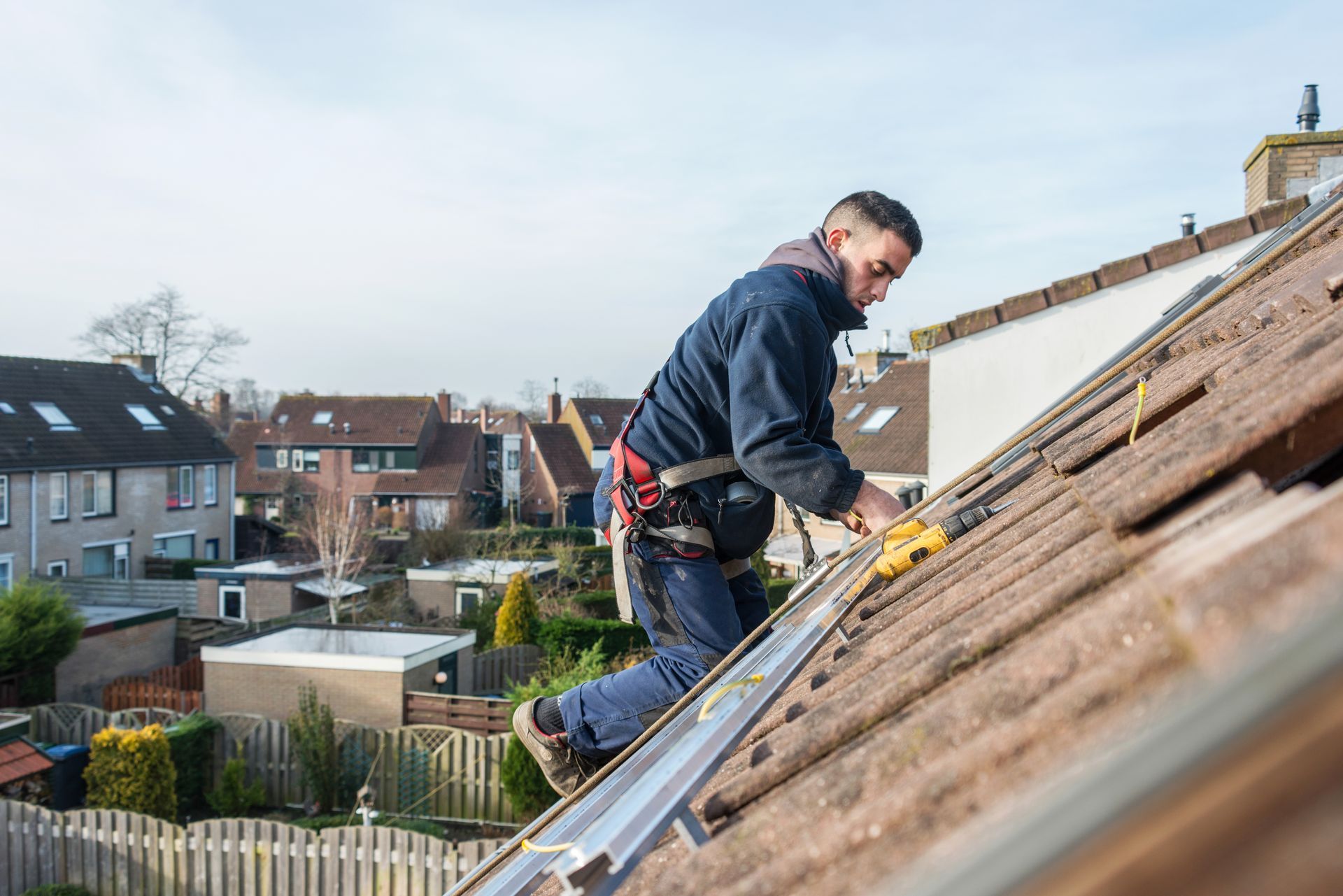 Roofer kneels on a sloped roof, working with tools on shingles in a suburban neighborhood.