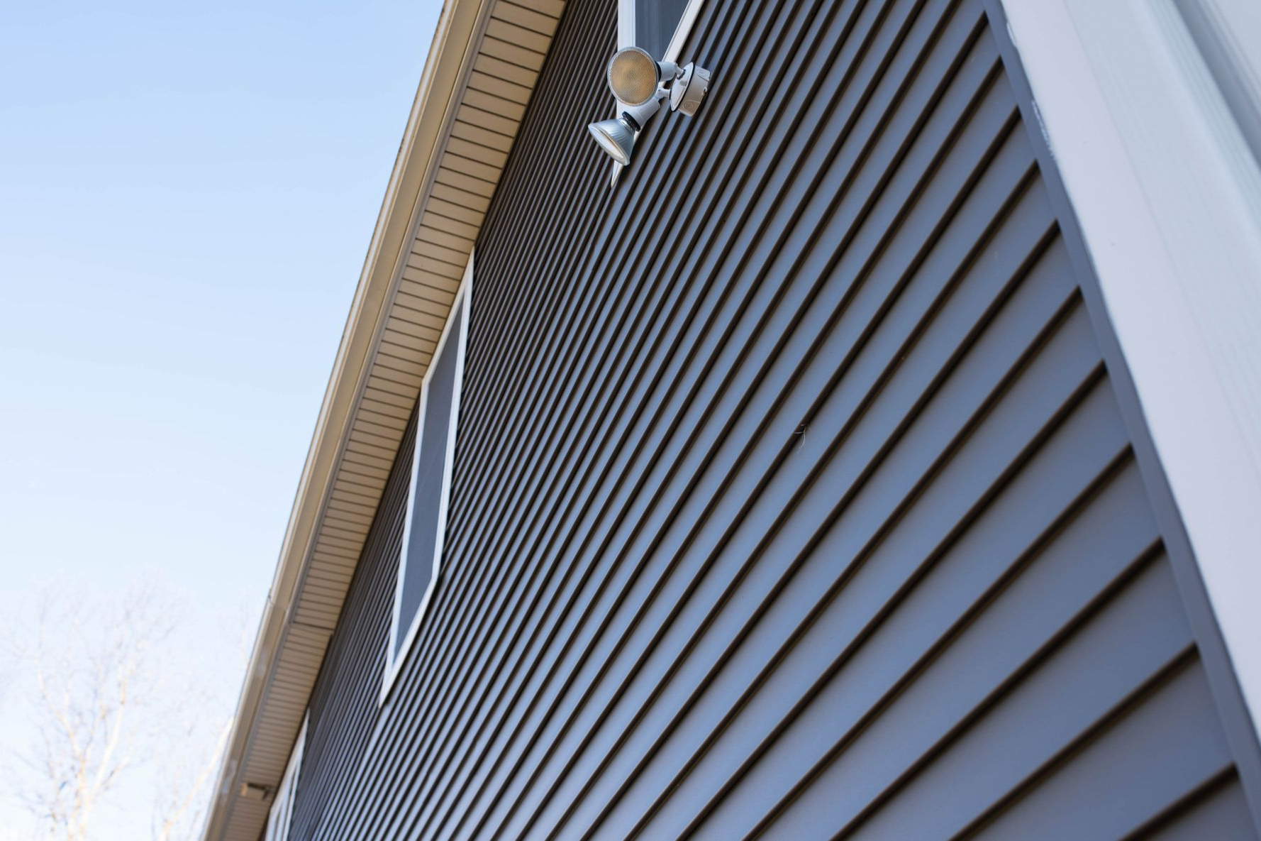 Dark gray siding on a building with a white window and spotlights under a beige eave against a blue sky.