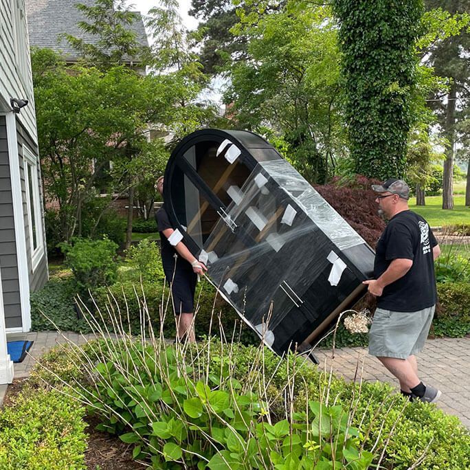 Two people carrying a large black curved object, likely furniture, outdoors near a house with landscaping.