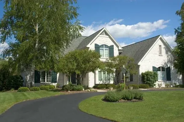 White brick house with green shutters, a curved driveway, and lawn under a blue sky