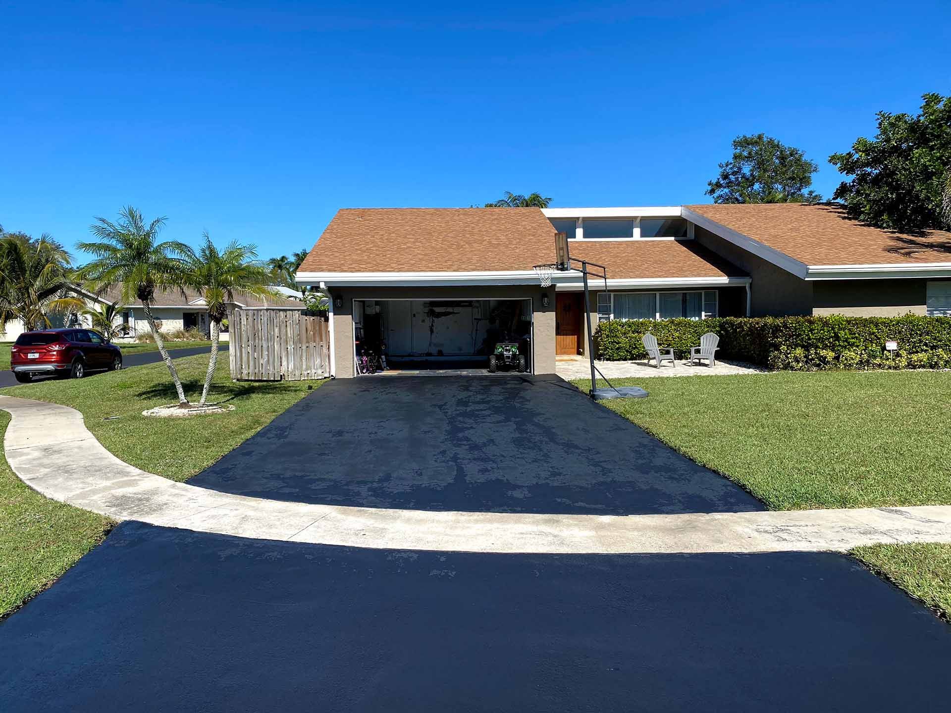 House with black asphalt driveway, lawn, palm trees, and clear blue sky
