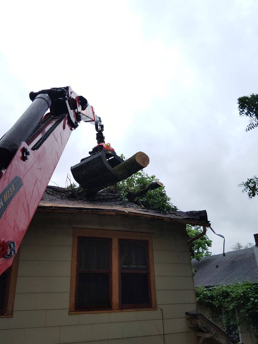 A red crane removing a tree limb from a house roof, under a cloudy sky.