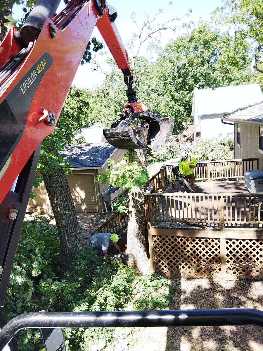 An orange excavator arm with a grapple removes tree branches near a wooden deck and house.