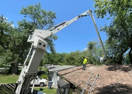 A worker in a yellow shirt stands on a residential roof, assisted by an elevated boom lift clearing nearby tree branches.