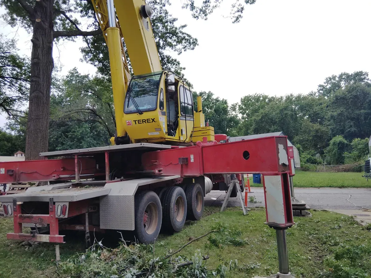 A yellow mobile crane with extended red outriggers sits on a grassy lot near trees and a road.