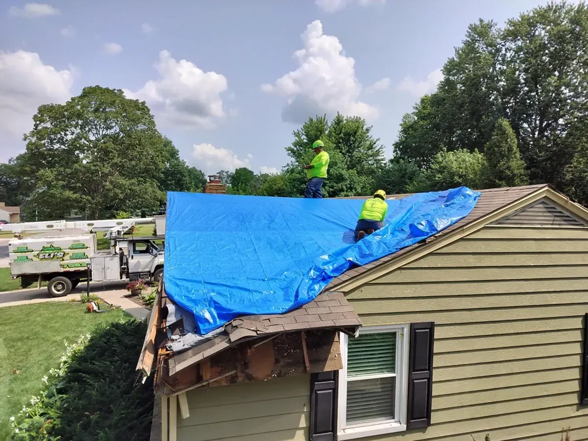 Two workers in high-visibility vests secure a large blue tarp over the damaged roof of a house with green siding.