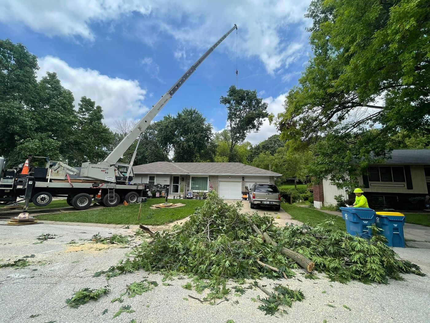 Tree removal service using a crane near a house. A worker in safety gear is collecting branches on a residential street.