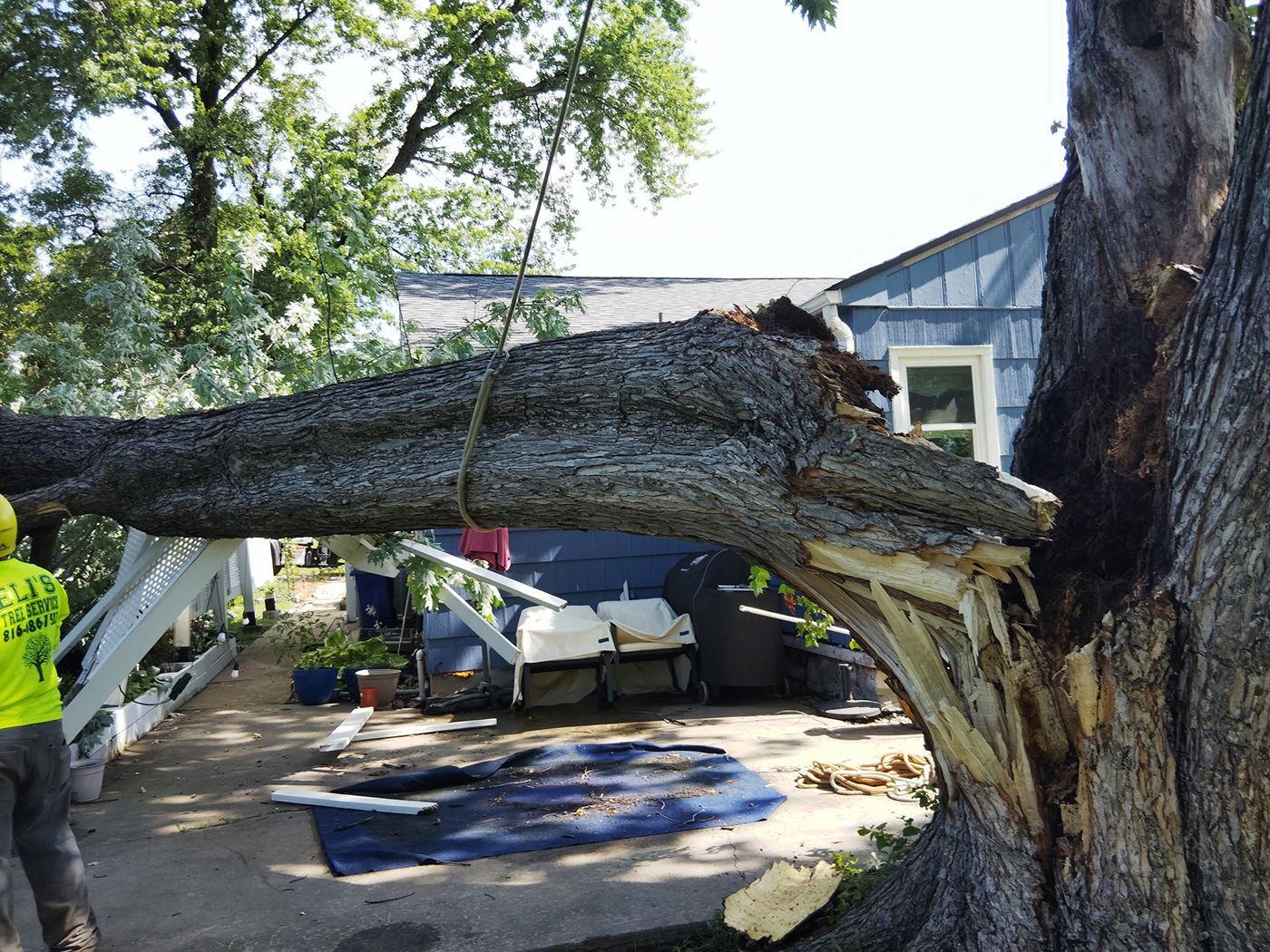 A large tree branch has fallen onto a home. A person in a neon shirt stands by a walkway.