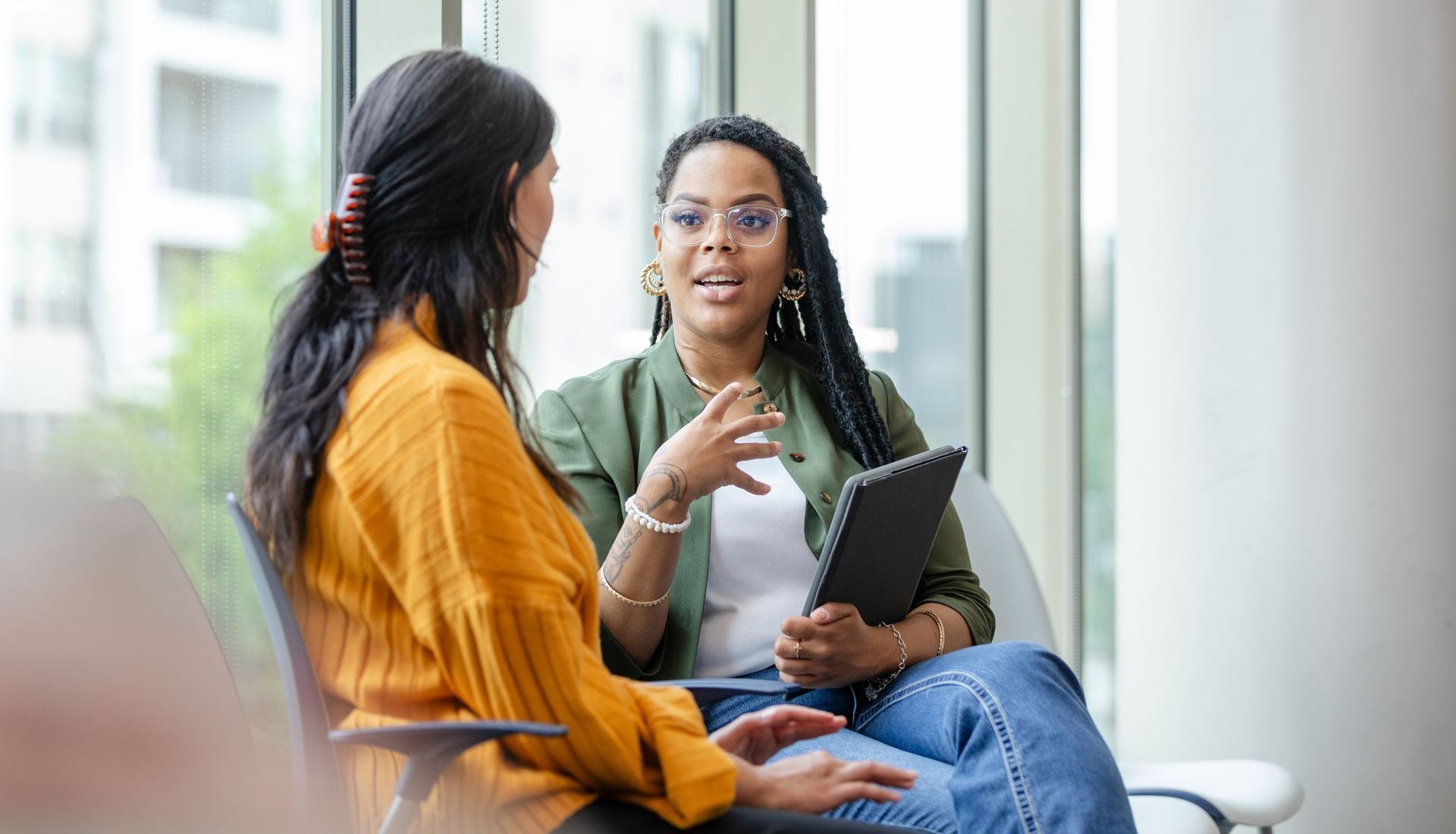 Two women are sitting in chairs talking to each other while holding a tablet.
