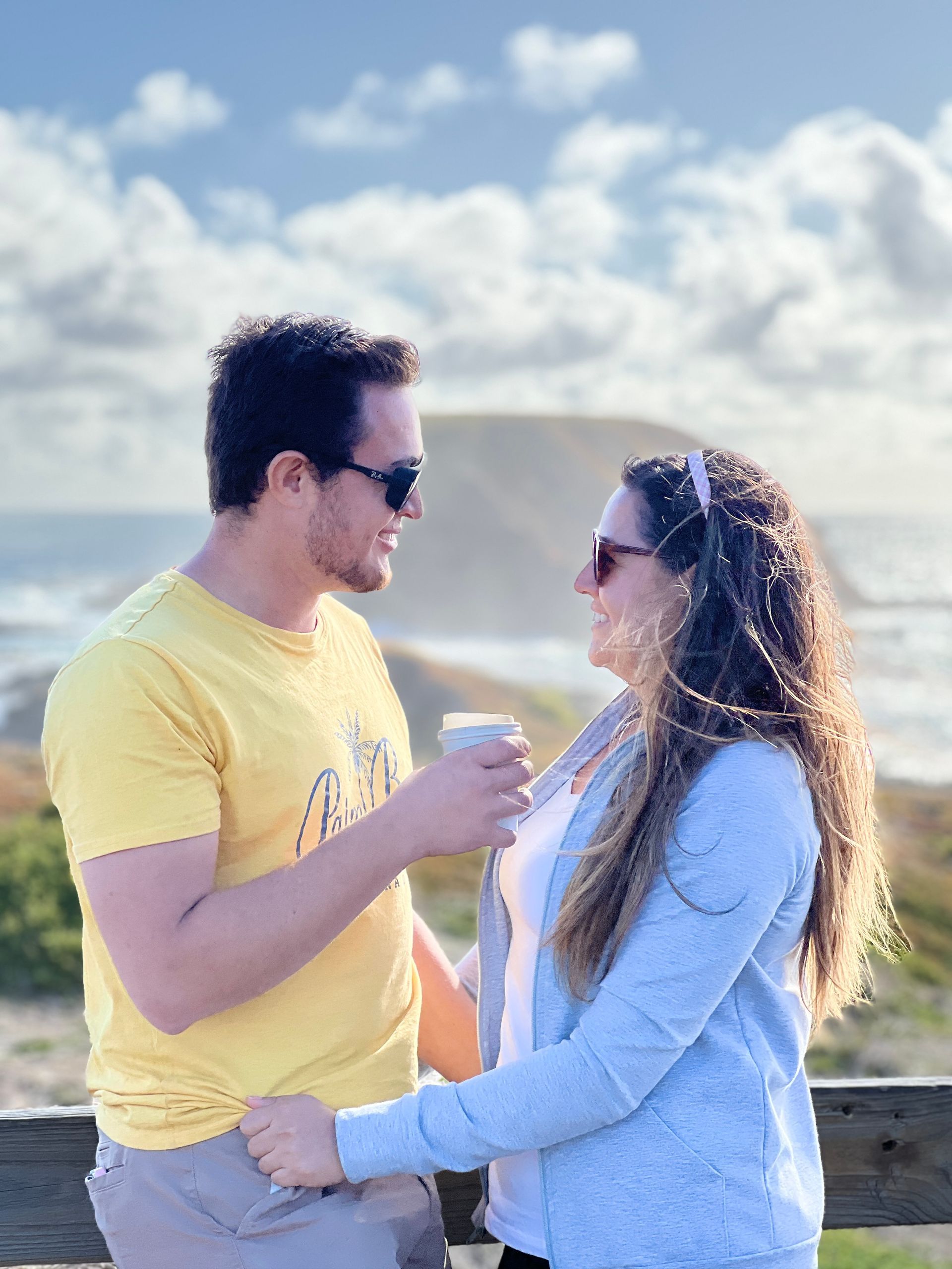 Pessoa segurando uma garrafa de água azul com adesivos, ao ar livre, com árvores e montanhas ao fundo.