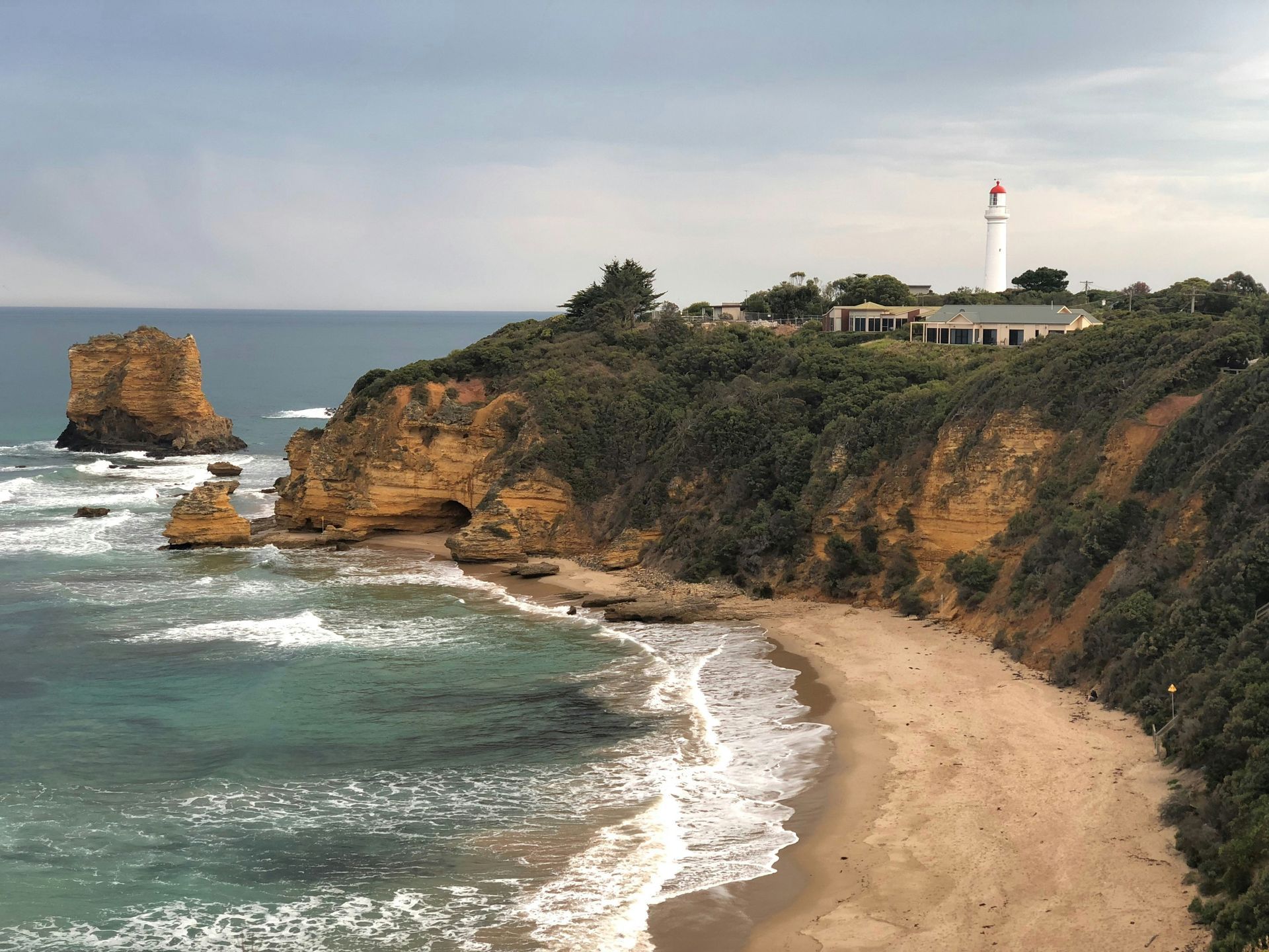 Vista costeira: farol em um penhasco, praia e oceano com uma formação rochosa. Céu nublado.