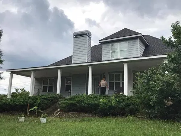 A person stands on the porch of a gray, two-story house with a chimney and wrap-around porch against a cloudy sky.