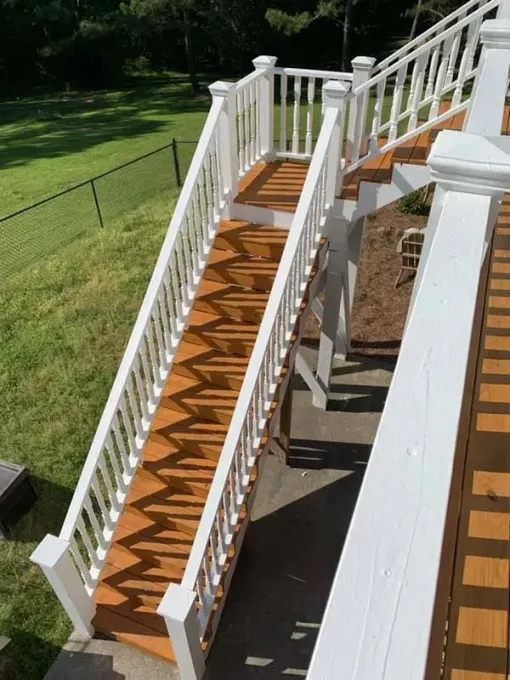 Wooden outdoor stairs with white railings leading down from a deck to a concrete patio and a grassy yard.