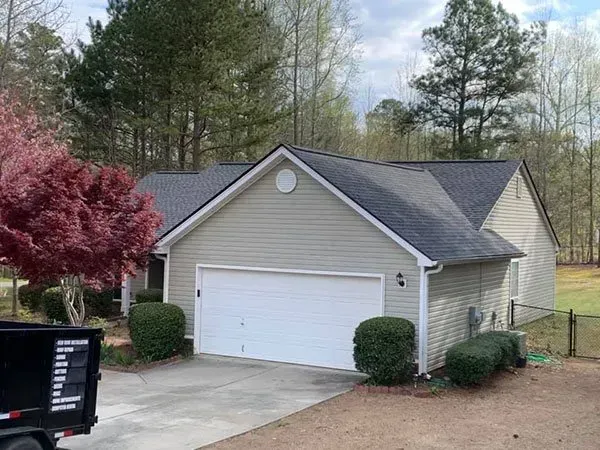 A tan single-story house with a white garage door, dark shingled roof, and a small red tree in the front yard.