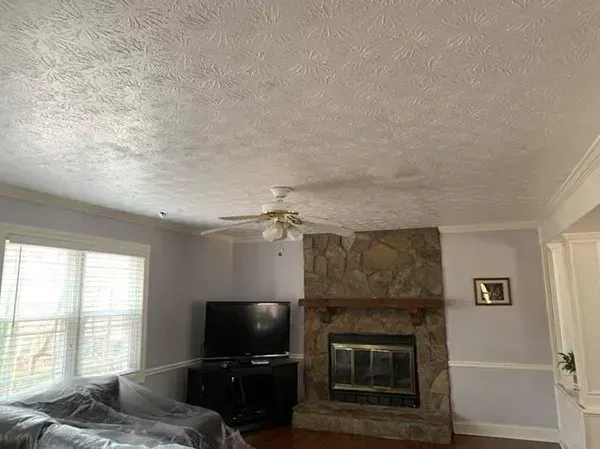 A living room with a stone fireplace, a black TV stand, a ceiling fan, and window blinds.