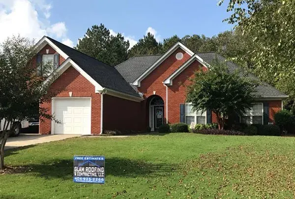 A red brick suburban house with a gray roof and a single-car garage, with a roofing contractor sign in the front lawn.