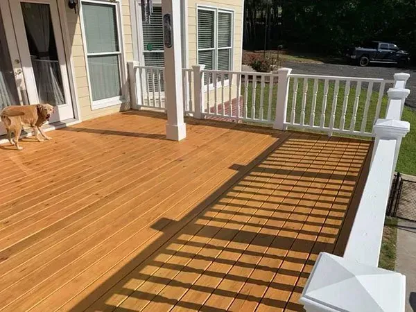 A light brown wooden deck attached to a house with white railings and a golden dog standing near the glass door.
