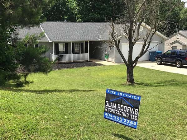 A single-story gray house with a front porch and a blue truck parked in the driveway, with a roofing business yard sign.