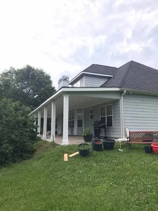 A house with light siding and a dark shingled roof features a covered patio with white columns, overlooking a grassy lawn.