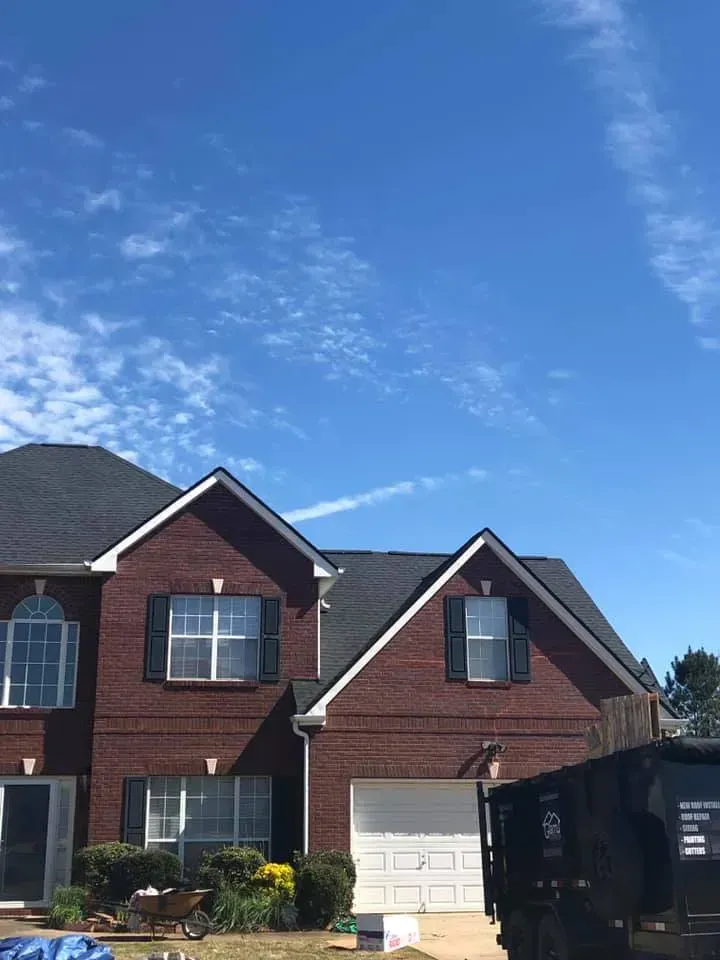A two-story brick house with a new dark shingle roof, featuring a garage and a large dumpster in the front yard.