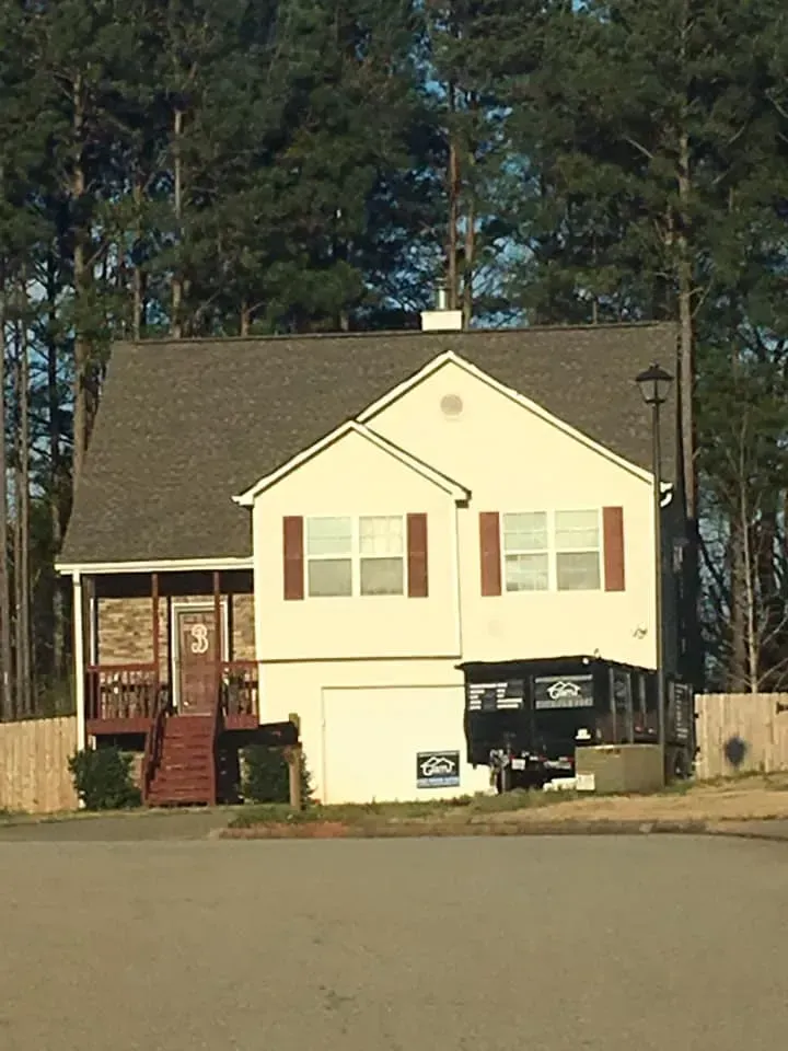 A light-colored two-story house with a wooden porch, a front-facing garage, and a black dumpster in the driveway.