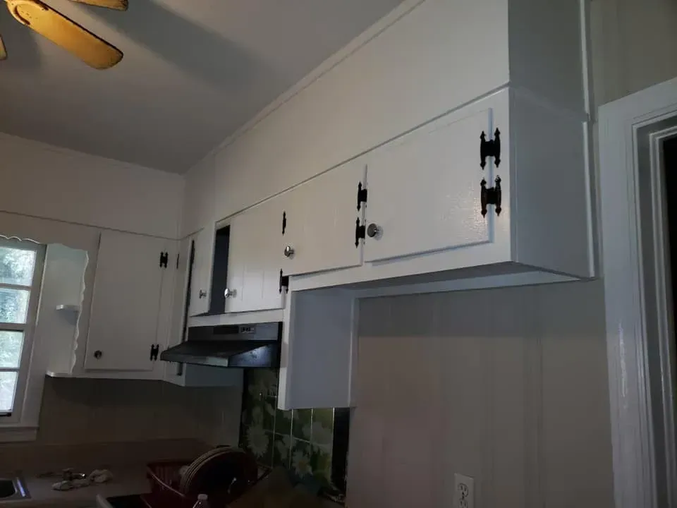 White kitchen cabinets with black exterior hinges mounted above a range hood and wall in a kitchen.