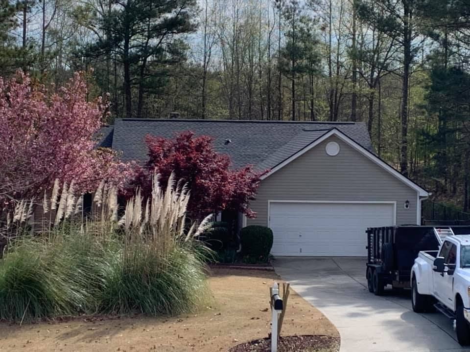 A beige house with a garage and a driveway containing a truck and a trailer, surrounded by trees and decorative bushes.