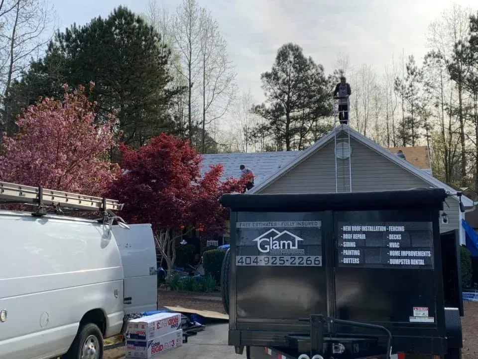 Workers on a residential roof with a company trailer parked in the driveway during a home improvement project.