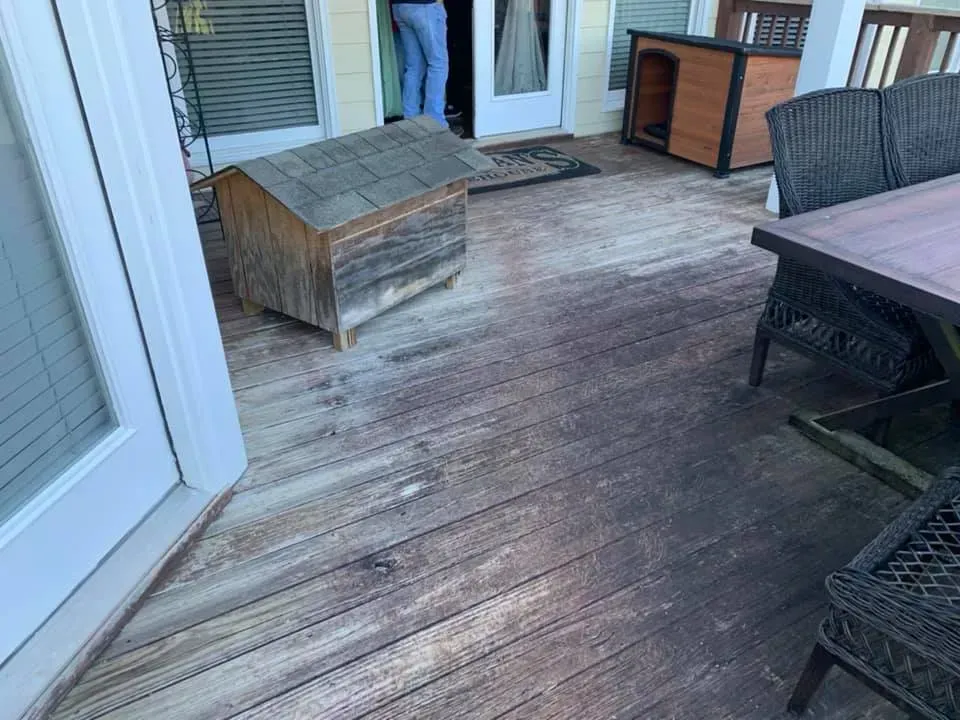 A weathered wooden deck showing a patch of lighter, cleaned wood next to darker, dirty planks, with pet houses nearby.