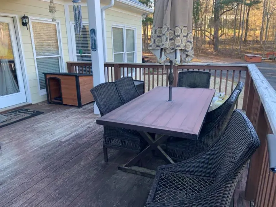 A wooden patio deck featuring a dark wood dining table with wicker chairs, an umbrella, and a small wooden dog house.