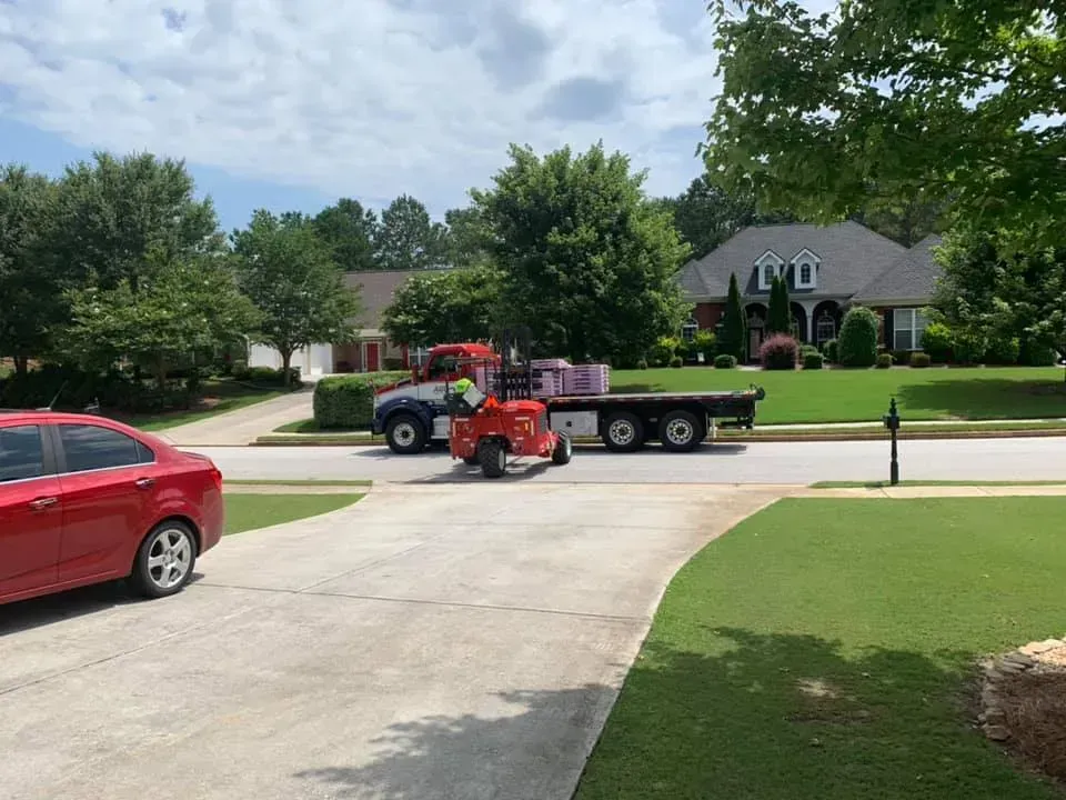 A flatbed delivery truck is parked on a suburban street, unloading pallets of building materials with a red forklift.