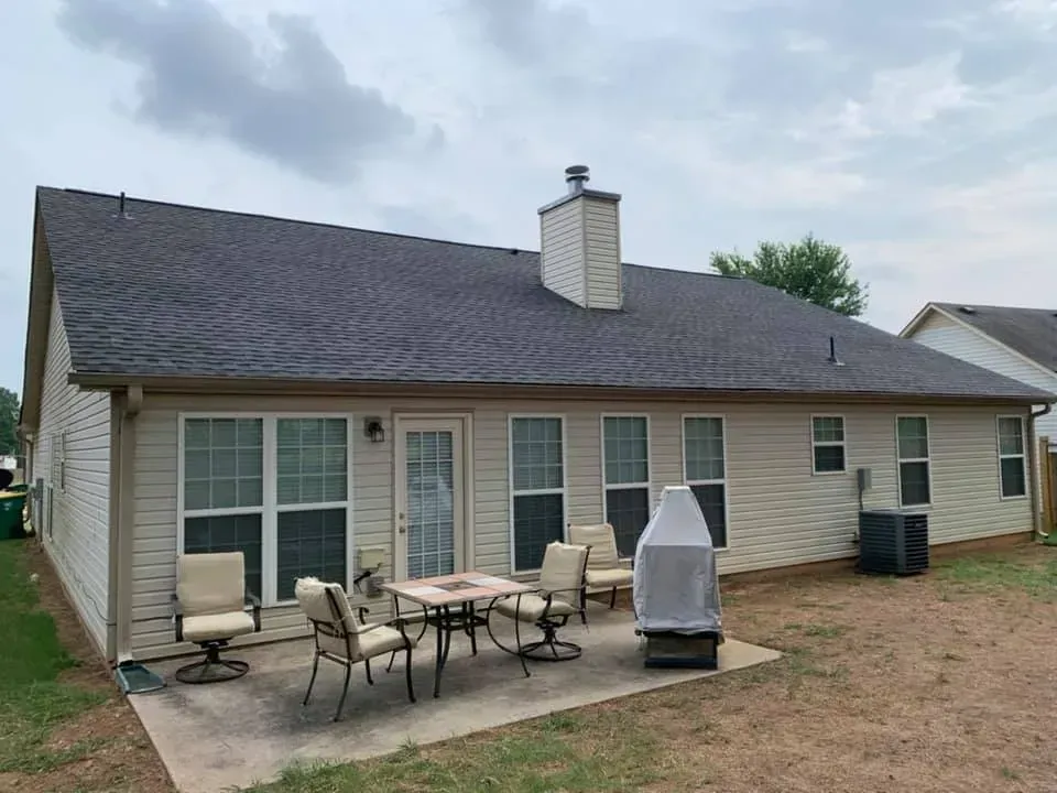 A beige vinyl-sided house with a dark shingled roof, a patio with chairs and a table, and a covered grill in the yard.