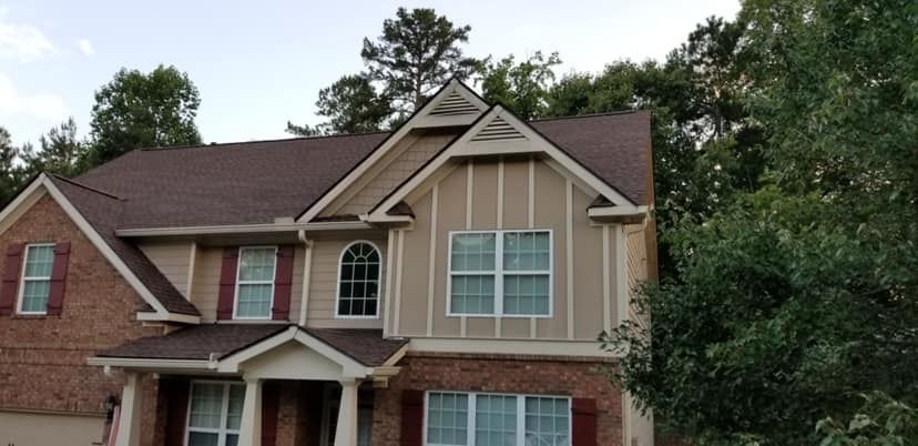 A two-story suburban house with a brick and beige siding facade, multiple gables, and dark shingles, surrounded by trees.