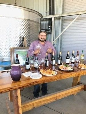 Man holding wine glass at a tasting table with various wine bottles, food, and a large tank in background.