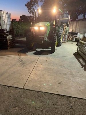 John Deere tractor with lights on, parked on concrete pad with a person standing nearby at dusk.