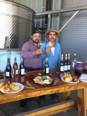 Two men toasting with wine glasses, standing behind a table with wine bottles and food.