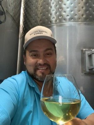 Man holding wine glass, smiling in a winery with stainless steel tanks in background. Wearing a hat and blue shirt.