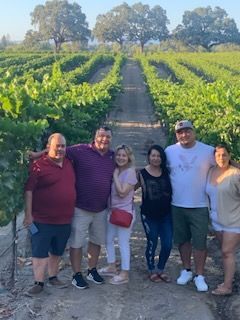 Group of six people posing for a photo in a vineyard, with rows of vines and trees in the background.