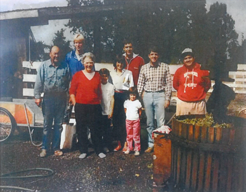 Group of people near a wooden press and cart outdoors. Some are smiling, likely a harvest celebration.