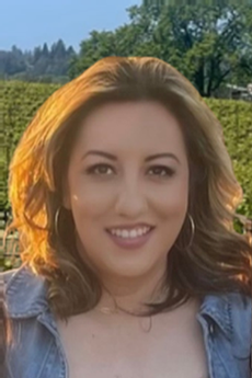 Woman smiling in front of a vineyard, wearing a denim shirt and hoop earrings.