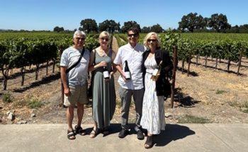 Four people stand in a vineyard, holding wine bottles. Sunny day; green vines in rows.