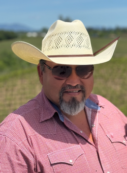 Man wearing a straw cowboy hat and sunglasses smiles outdoors, in a pink shirt.