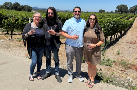 Four people holding wine glasses in a vineyard on a sunny day.
