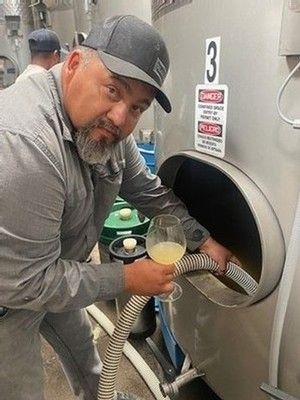 Man in grey holding wine glass, sampling from a metal tank. Inside a winery, by a warning sign.