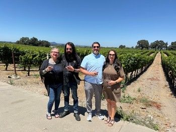 Four people standing in a vineyard, holding wine glasses, under a blue sky.