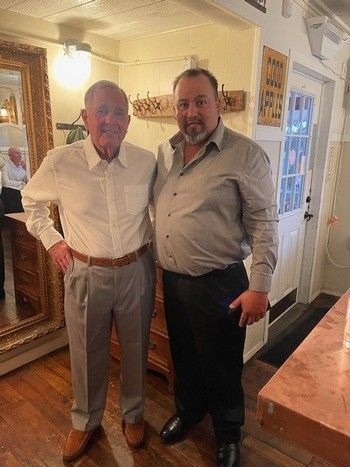 Two men standing inside a room. One man wears a white shirt and gray pants. The other man wears a gray shirt and black pants.