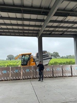 Man stands in front of boxes, with machinery and field in the background under a metal structure.